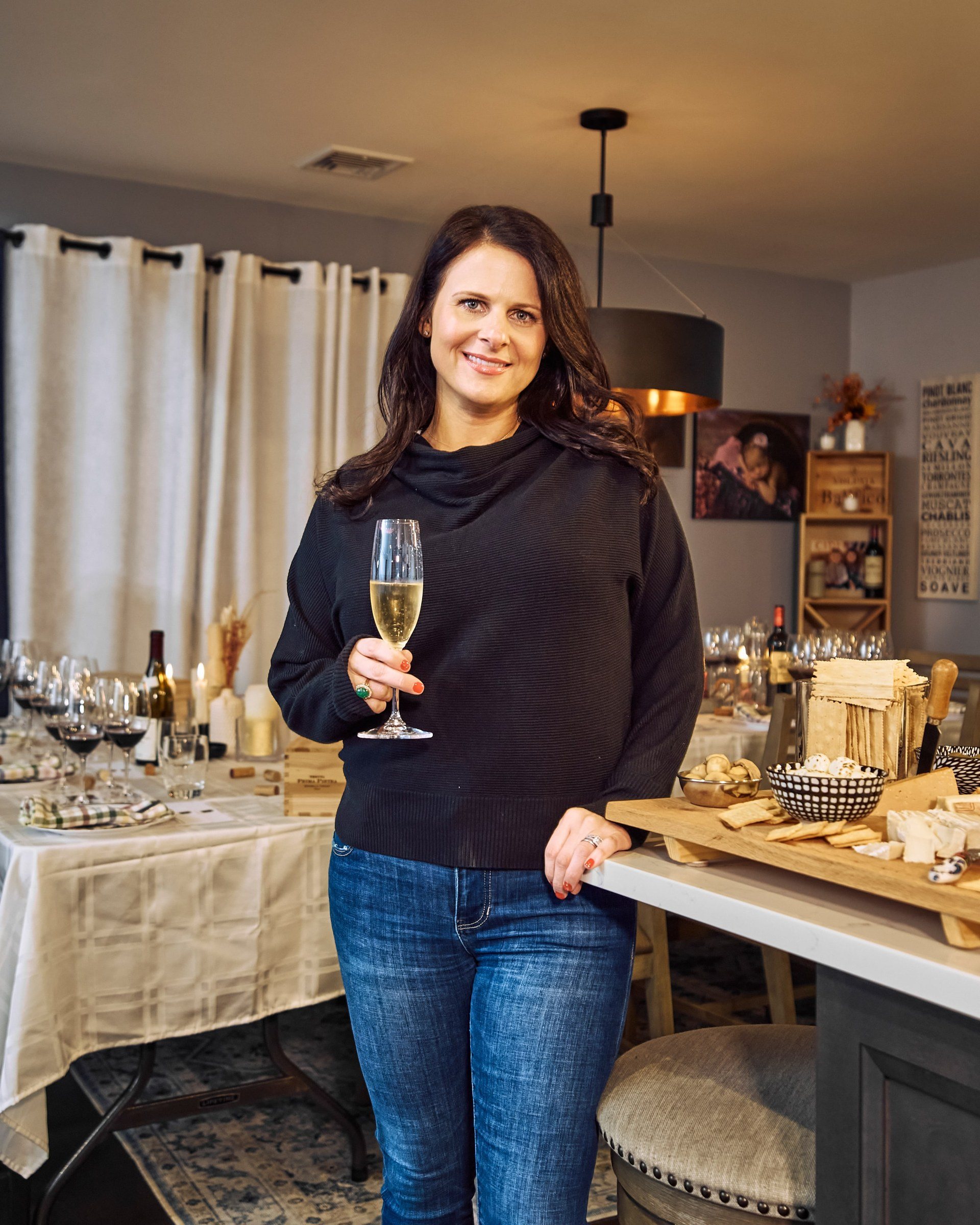 Alicia Abaté in a dark jumper, holding a glass of sparkling wine beside the set table.