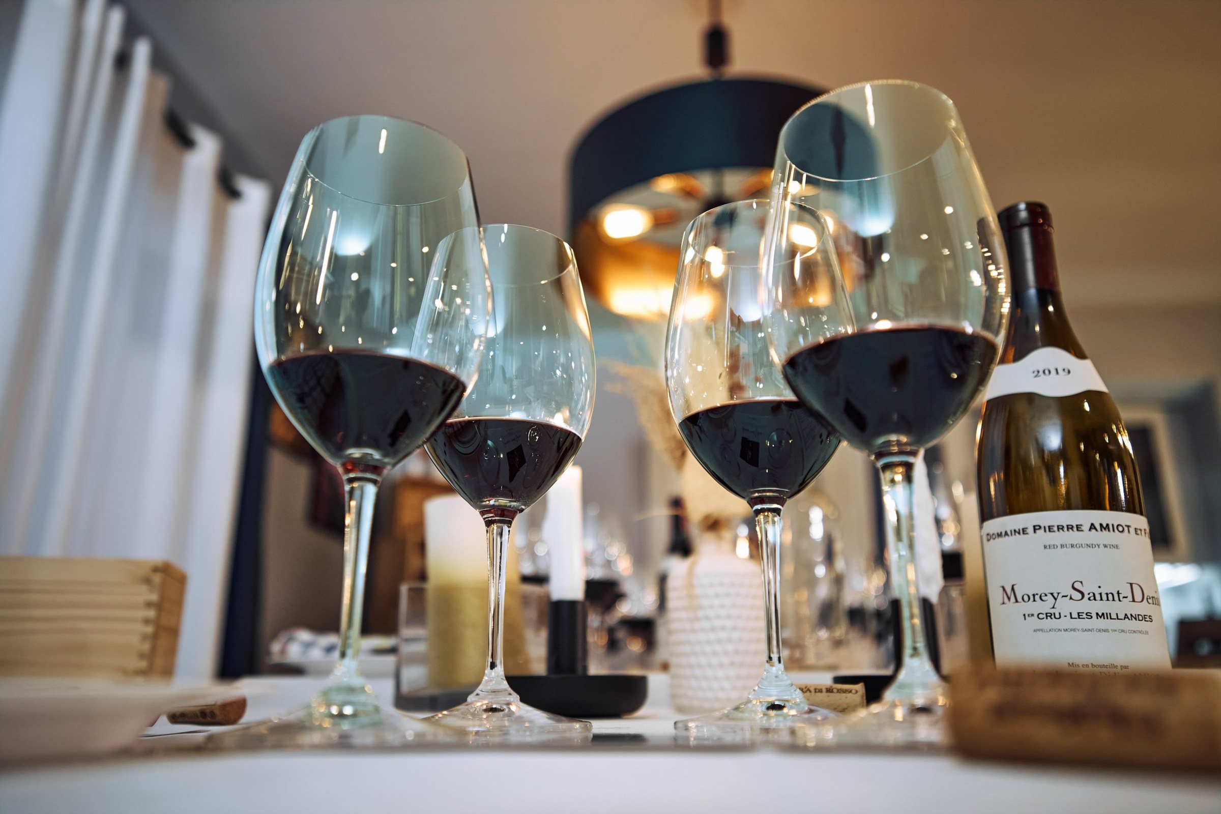Glasses of red wine and a bottle of burgundy on the table under a copper pendant.
