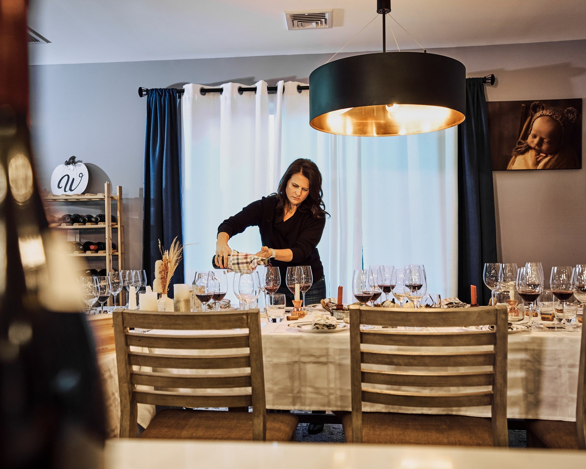 Alicia setting glasses along a long table under a black-and-copper pendant light before guests arrive.