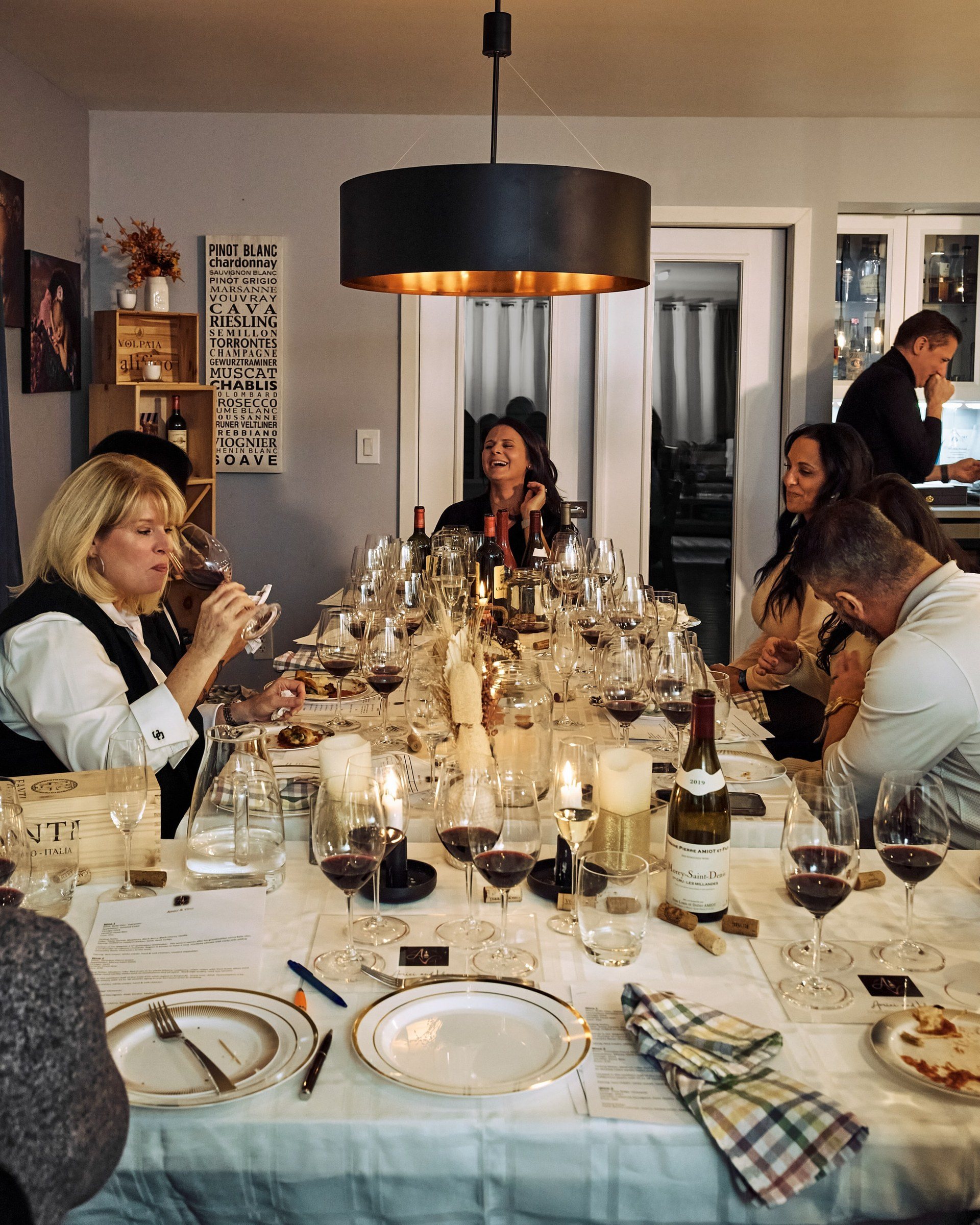 Guests round a long candlelit table with glasses of red wine, tasting notes at each place.