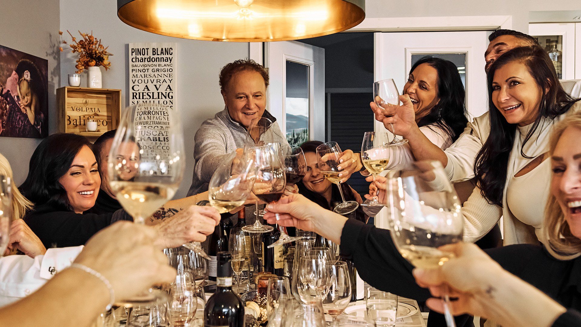 Guests round a long table raise glasses of white and red wine towards each other, laughing, under a copper pendant light.