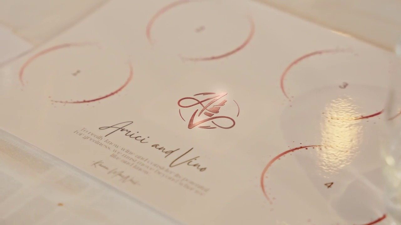 A white tasting placemat with numbered circles for glasses, the rose-copper monogram and Amici and Vino in script.