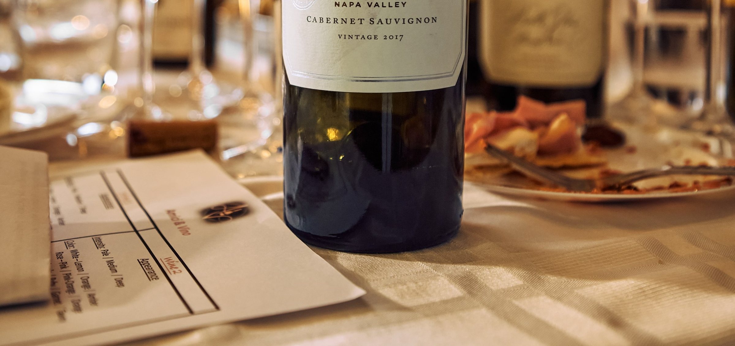A tasting sheet beside a bottle: the monogram and Amici & Vino in rose copper above a grid for appearance, intensity and colour.