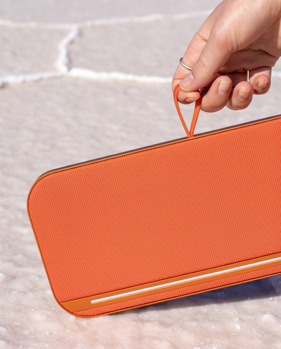 A hand lifting the orange AMP speaker by its strap loop above a white salt flat