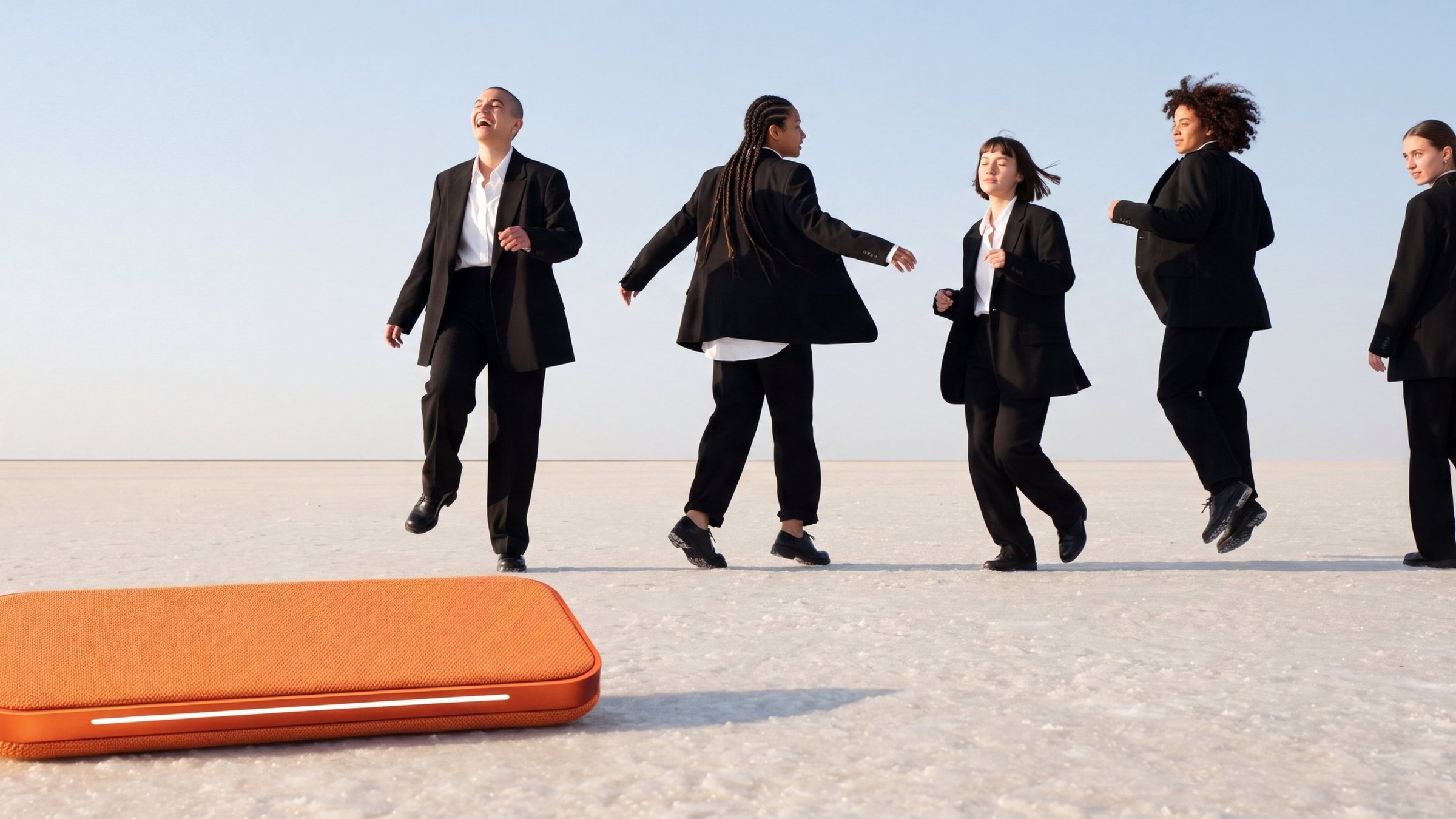 Five people in black suits dancing across a white salt flat, far from an orange speaker resting in the foreground