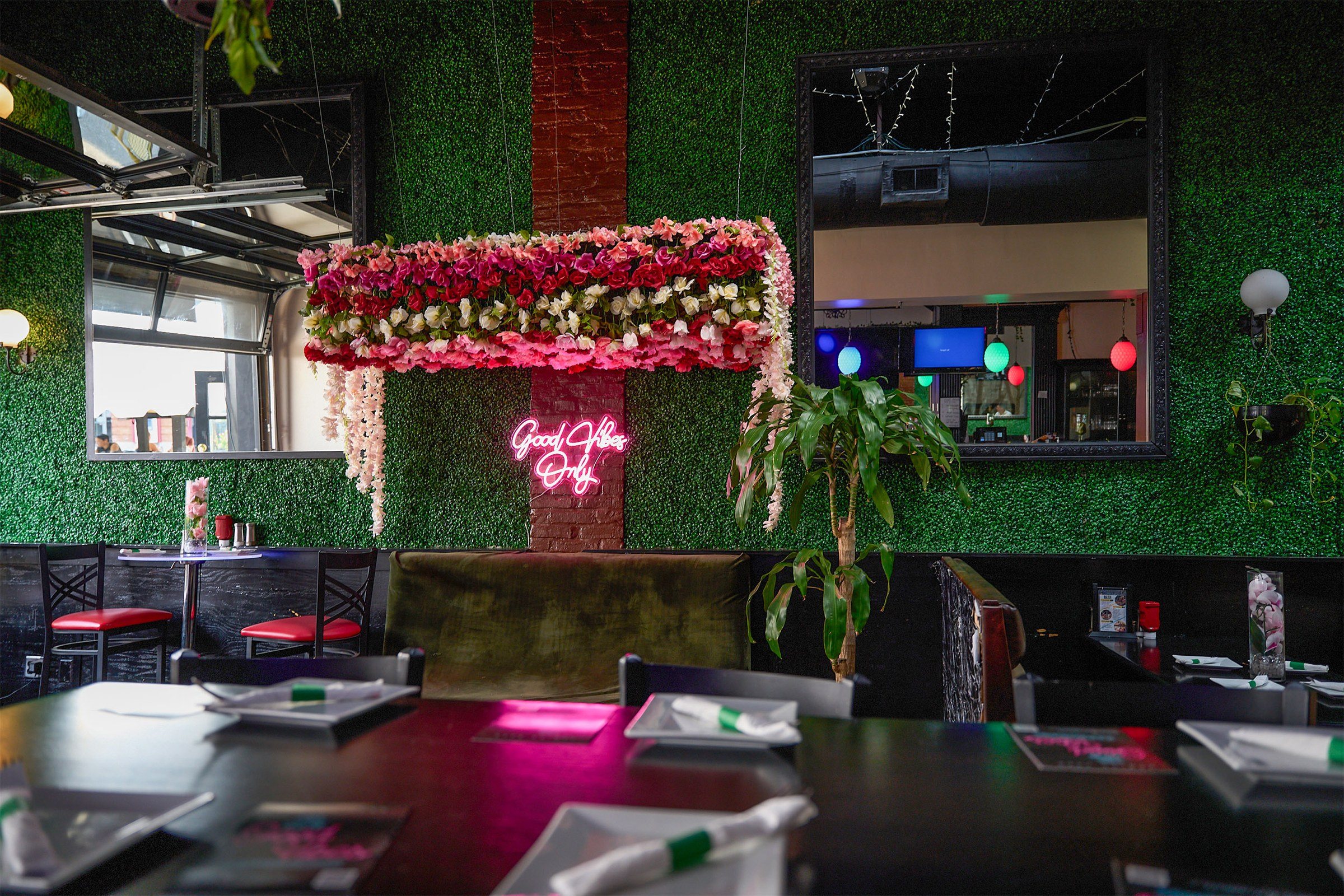 Inside the old Good Vibes: a wall of green hedge with a canopy of pink flowers and a pink Good Vibes Only neon, booths and tables in front.