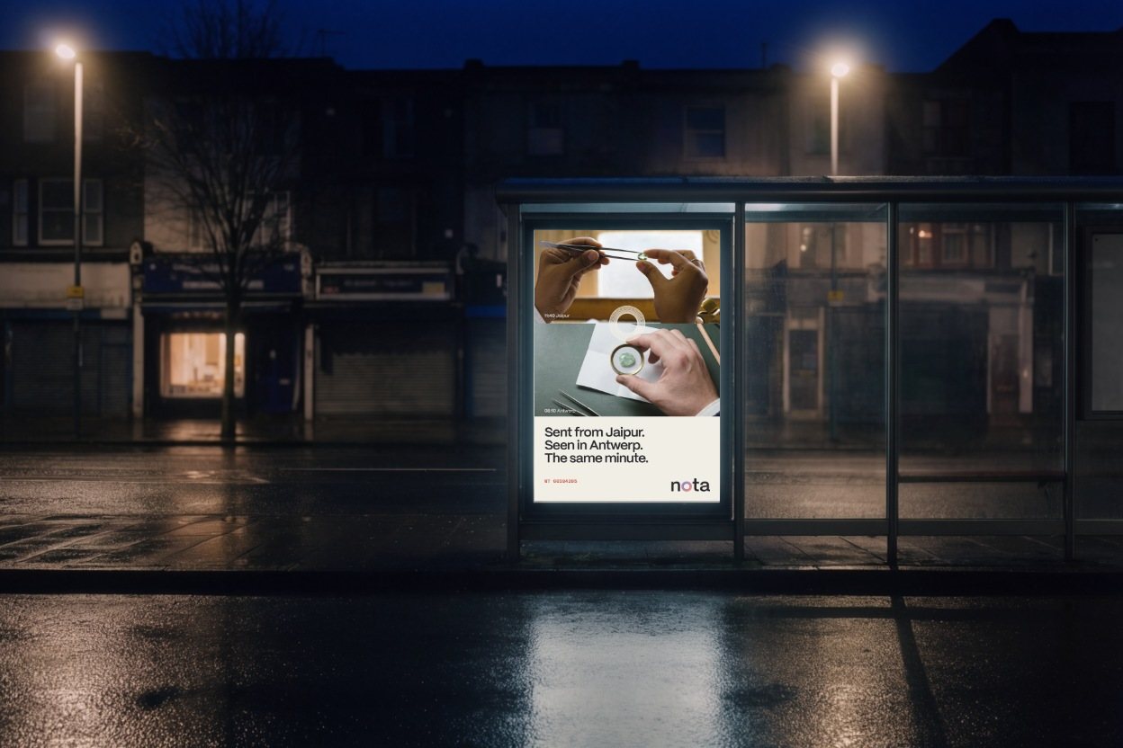 A lit poster in an Antwerp bus shelter at night: the Jaipur stone above the seam, the Antwerp loupe below, Sent from Jaipur. Seen in Antwerp. The same minute.