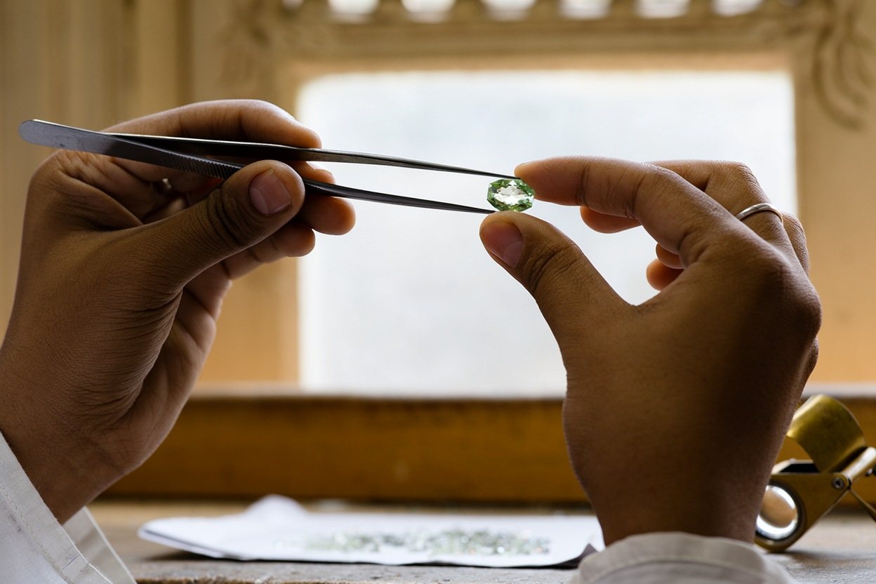Hands in a Jaipur workshop hold a pale green stone up to the window with tweezers.
