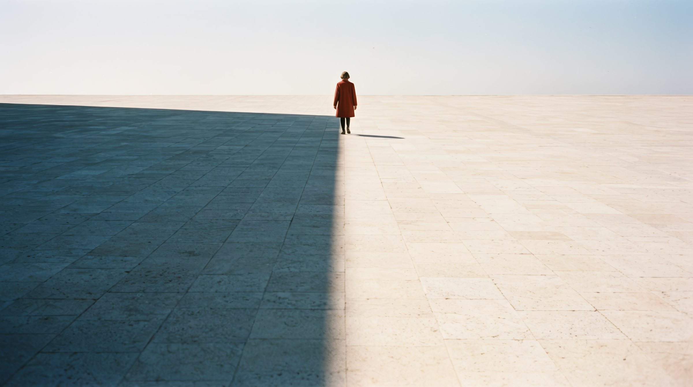 A vast stone plaza split by a straight shadow edge; a small figure in a rust red coat steps from the shadow into the sun