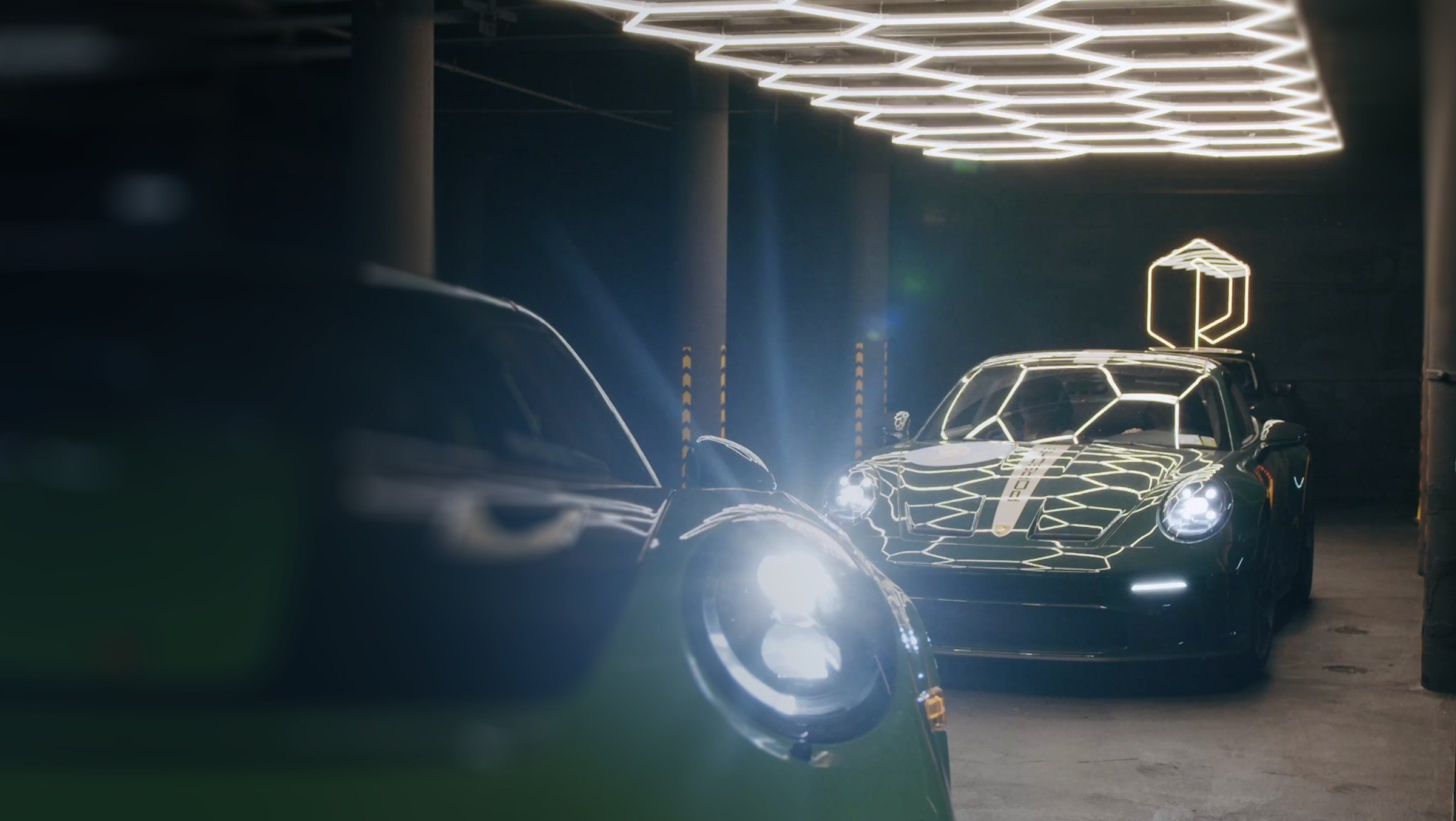A black Porsche 911 in the Manhattan garage, its body traced in lines of reflected hexagonal light, a neon hexagon mark above it