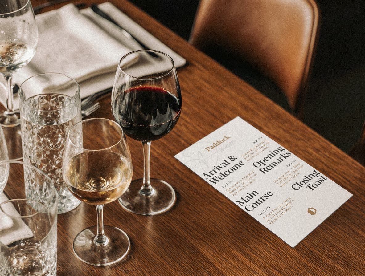 A Paddock dinner menu on a wooden table between wine glasses: Arrival and Welcome, Opening Remarks, Main Course, Closing Toast.