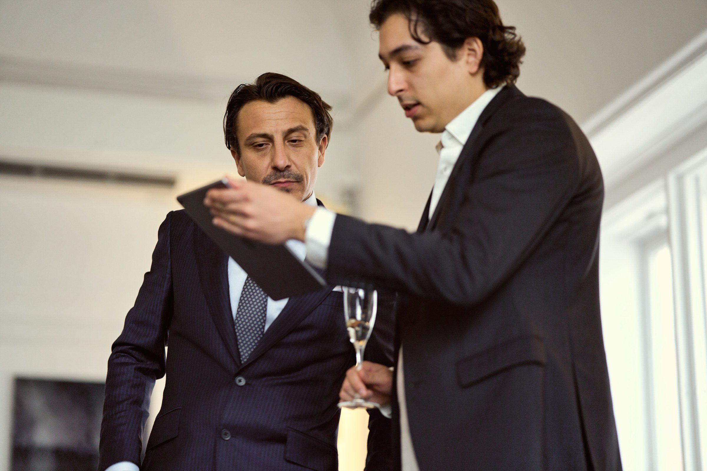 Two men in tailoring talking by a window, one holding a glass.