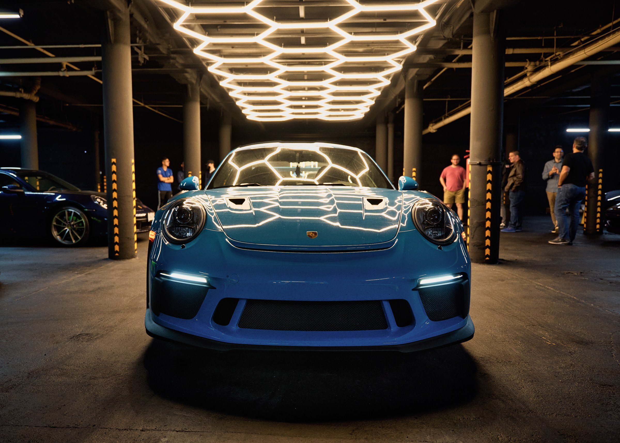 A blue Porsche 911 GT3 centred under the hexagonal light ceiling, guests behind it.