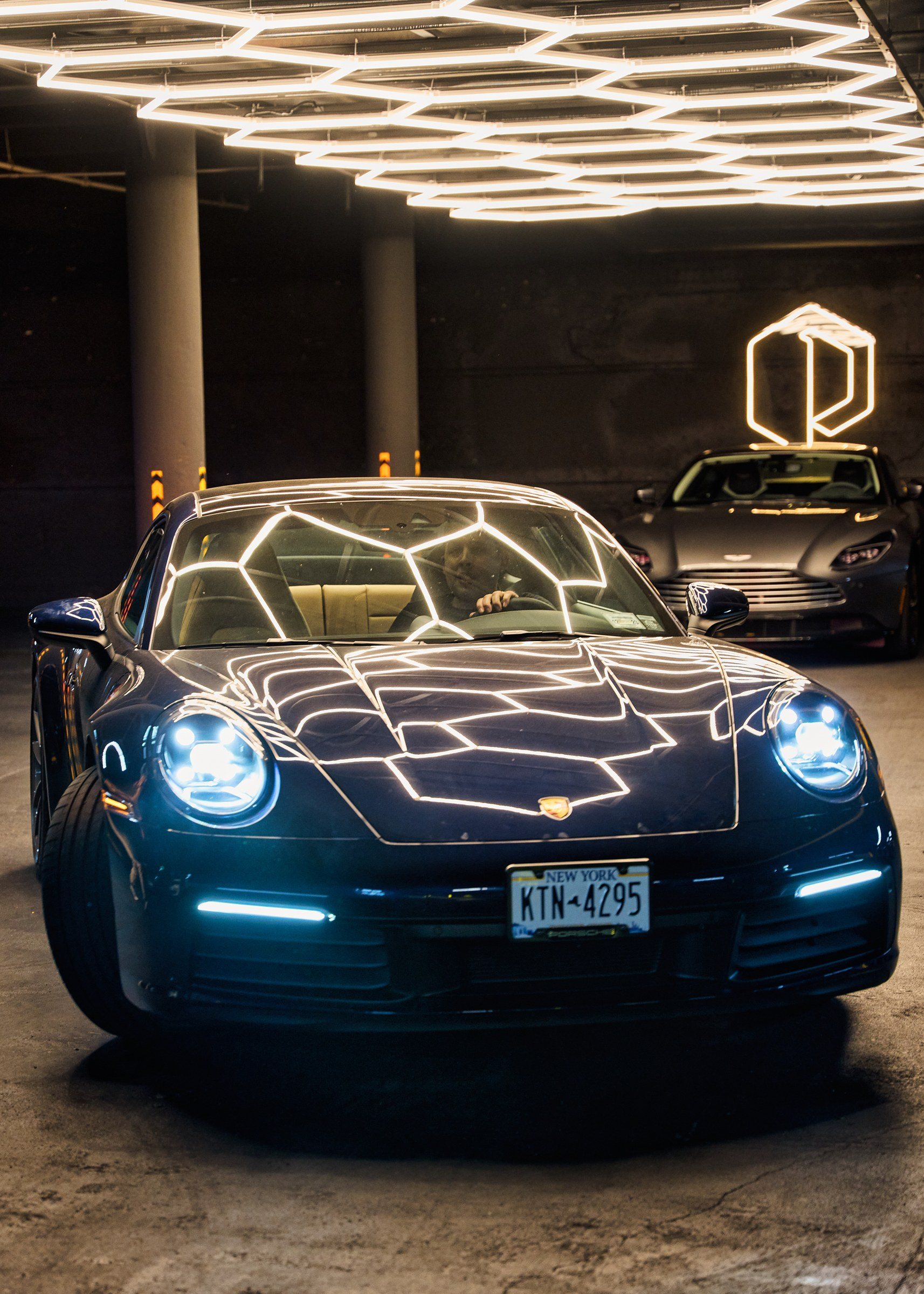A blue Porsche 911 under the hexagonal light ceiling, with the mark lit as a neon lightbox behind it.