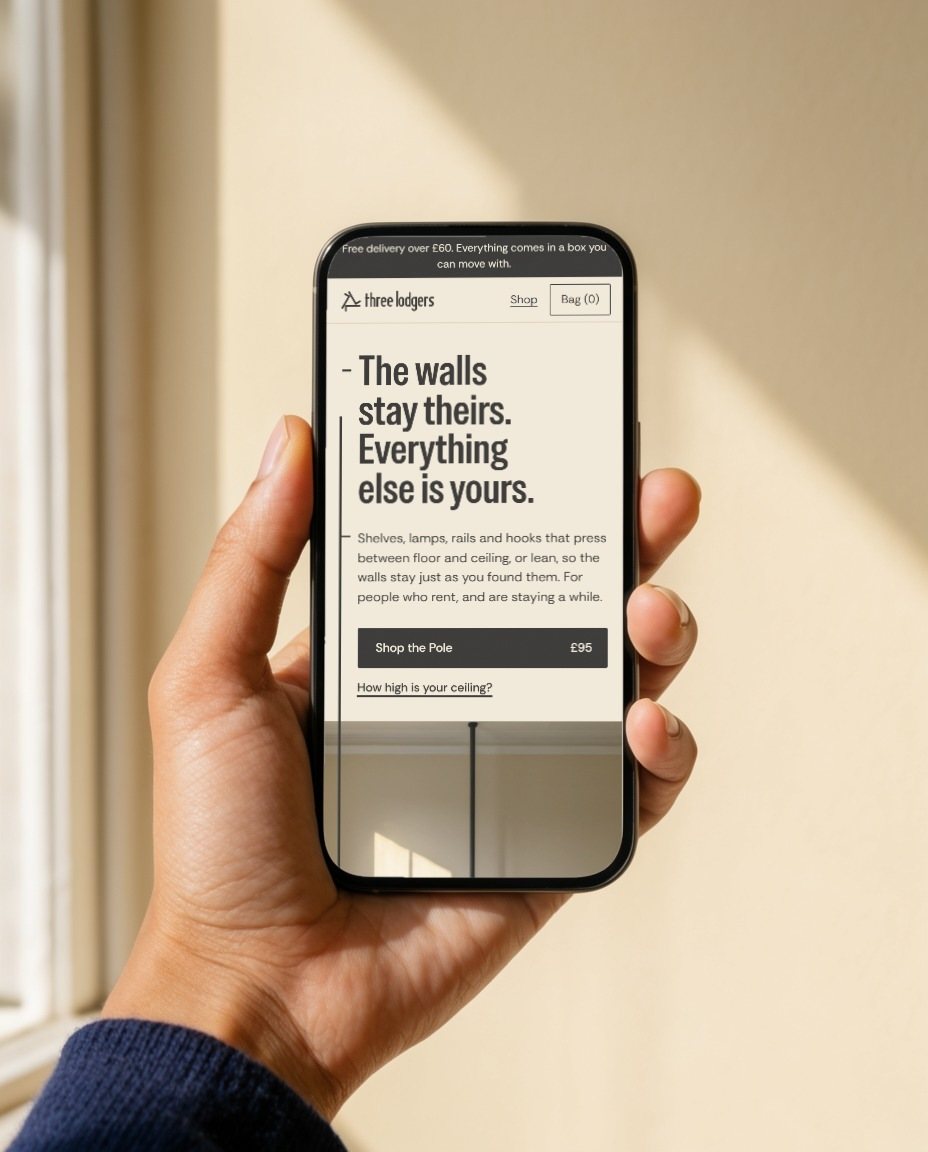 A hand holds a phone showing the Three Lodgers homepage against a sunlit wall.
